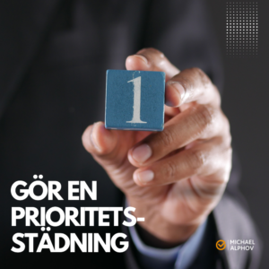 En person i kostym håller fram en blå träkloss med siffran 1, med texten "Gör en prioritetsstädning" i nedre vänstra hörnet och Michael Alphovs logotyp i nedre högra hörnet.